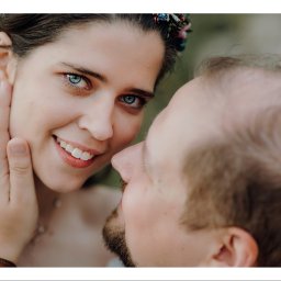 OTO STUDIO Sp. z o.o. - Uśmiechnięta kobieta z wiankiem na głowie i niebieskimi oczami, delikatnie dotykana po twarzy przez mężczyznę, zbliżenie.