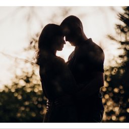 OTO STUDIO Sp. z o.o. - Silhouetted couple embracing at sunset, soft bokeh background, branches in foreground, logo in corner.