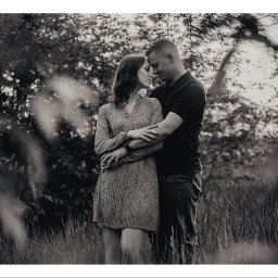 OTO STUDIO Sp. z o.o. - Czarno-białe zdjęcie zakochanej pary w objęciach na tle drzew i wysokiej trawy, delikatne światło słoneczne przebija przez liście.
