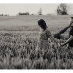 Czarno-białe zdjęcie pary trzymającej się za ręce, idącej przez pole zboża; kobieta w sukience w drobny wzór, mężczyzna w ciemnej koszulce.