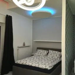 Nowoczesna sypialnia z łóżkiem w szarej ramie, wzorzystą pościelą, okrągłą lampą sufitową w kształcie planet i podświetlanym sufitem w kolorze niebieskim.