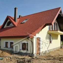 Żółty dom jednorodzinny z czerwoną dachówką, widoczny komin, mały balkon i sterta ziemi przed budynkiem.