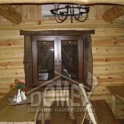 Wnętrze domu z bali z drewnianym stołem, krzesłami i oknem w drewnianej ramie, oświetlone żyrandolem z metalu, z widocznymi elementami instalacji klimatyzacyjnej pod sufitem.