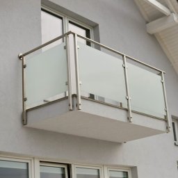 MGX Sp. z o.o. - Kwadratowy balkon z balustradą ze stali nierdzewnej i matowego szkła, zamontowany na szarej fasadzie domu z widocznymi oknami.