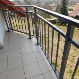 MGX Sp. z o.o. - Nowoczesna, szara balustrada metalowa na balkonie wyłożonym jasnymi płytkami, widok z góry na otoczenie z domami i drzewami.