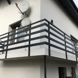 Nowoczesna, czarna balustrada metalowa z poziomymi prętami na balkonie budynku z białymi ścianami i dwoma antenami satelitarnymi.