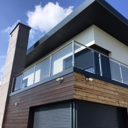 MGX Sp. z o.o. - Nowoczesny dom z drewnianą elewacją i szklanymi balustradami balkonowymi na tle błękitnego nieba z chmurami.