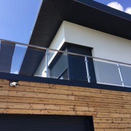 MGX Sp. z o.o. - Nowoczesna balustrada szklana z elementami ze stali nierdzewnej na balkonie w Bolęcinie, zamontowana na elewacji z drewnianą okładziną i widoczną kamerą monitoringu.