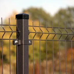 Fragment brązowego ogrodzenia panelowego z metalowym słupkiem i nasadką, widok z bliska na tle jesiennego krajobrazu.