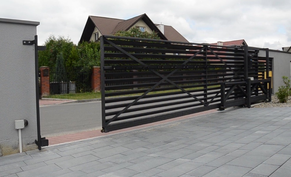 Czarna, przesuwna brama wjazdowa z poziomych profili, widoczny mechanizm automatycznego otwierania, szara kostka brukowa na podjeździe, dom z brązowym dachem w tle.