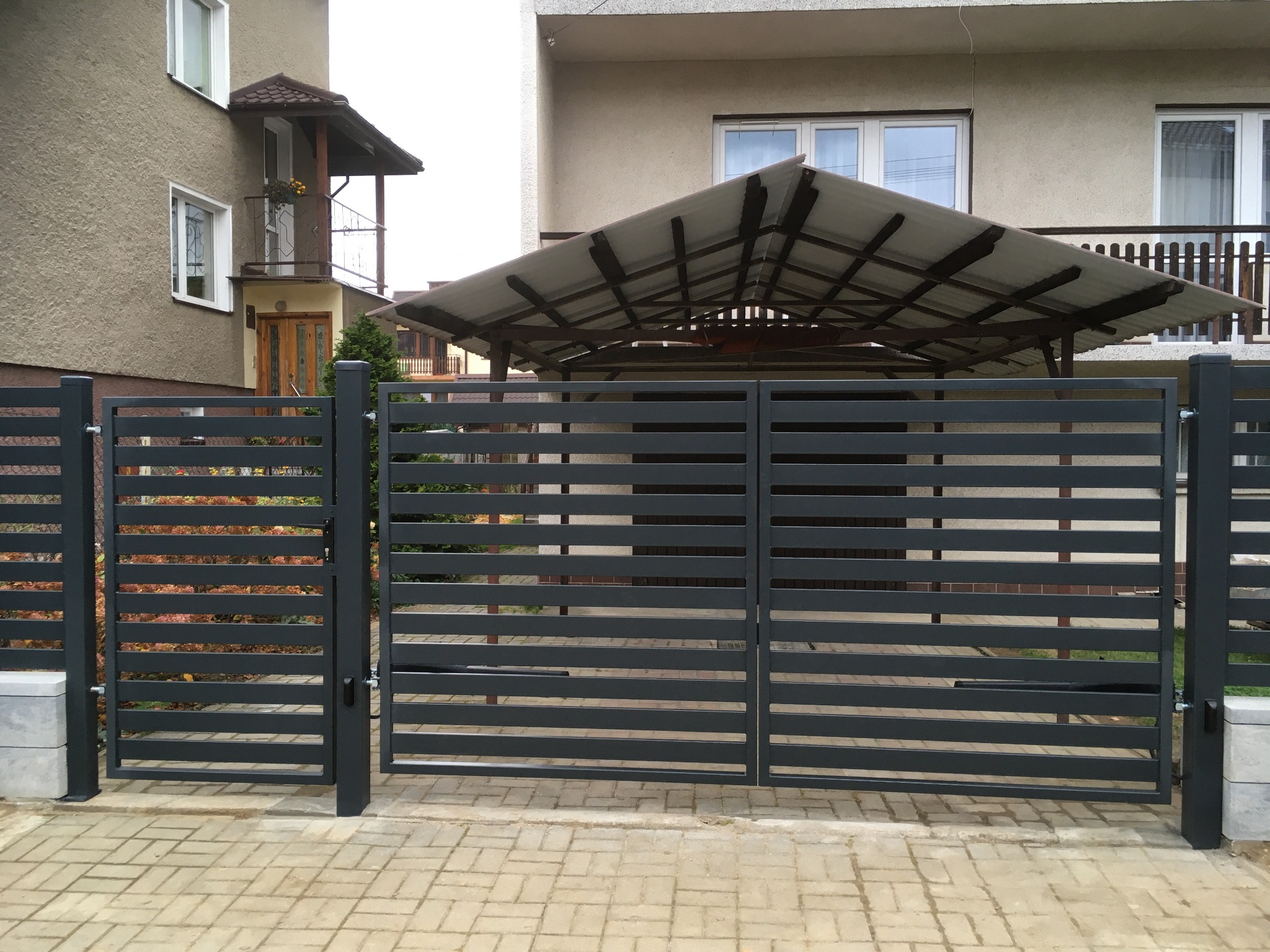 Nowoczesna, pozioma brama wjazdowa i furtka w kolorze antracytowym, z widoczną kostką brukową i budynkami w tle.