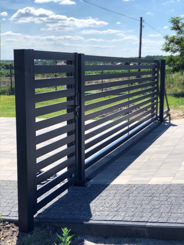 Nowoczesna, grafitowa brama wjazdowa z poziomymi panelami, widok z zewnątrz na tle zieleni i błękitnego nieba, z widocznym fragmentem podjazdu wyłożonego szarą kostką.