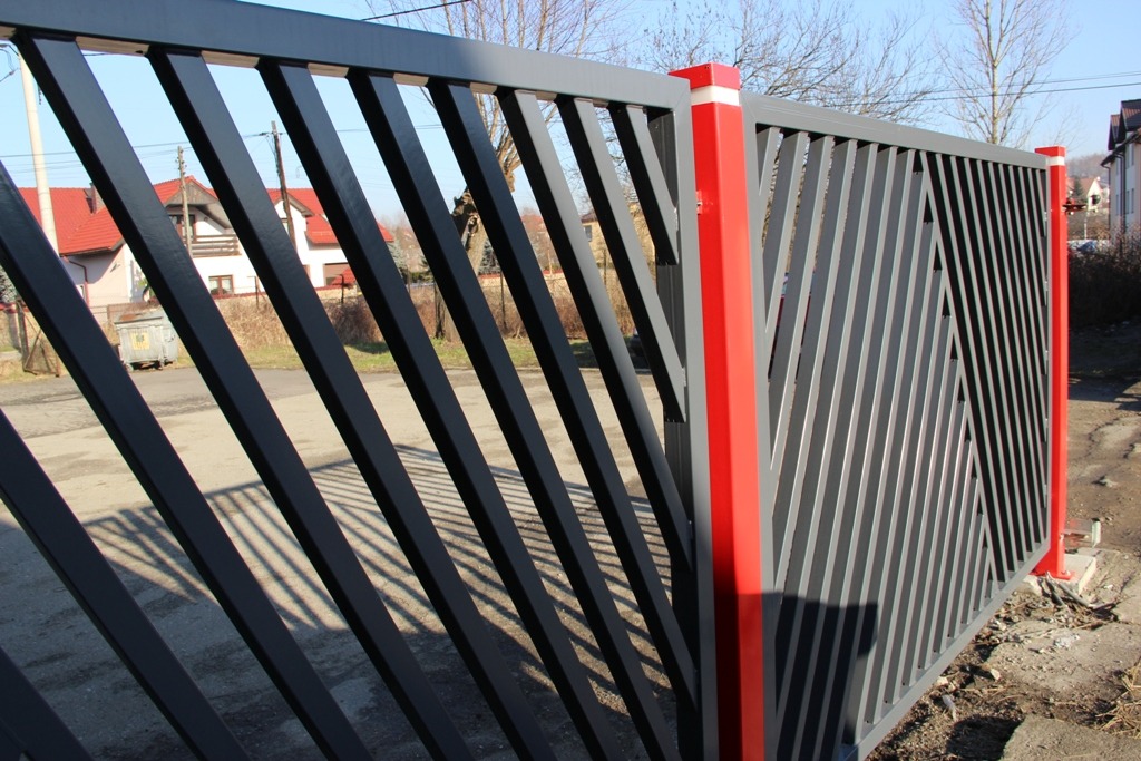 Nowoczesna brama wjazdowa z ukośnymi, metalowymi prętami w kolorze grafitowym, z czerwonymi słupkami, na tle ulicy i zabudowań.