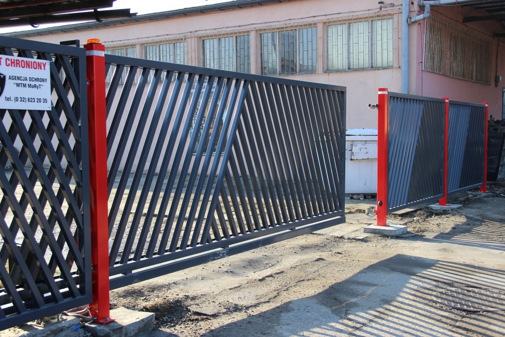 Otwarta przesuwna brama wjazdowa z czarnymi ukośnymi prętami i czerwonymi słupkami, na betonowym podłożu, z tabliczką informacyjną agencji ochrony.
