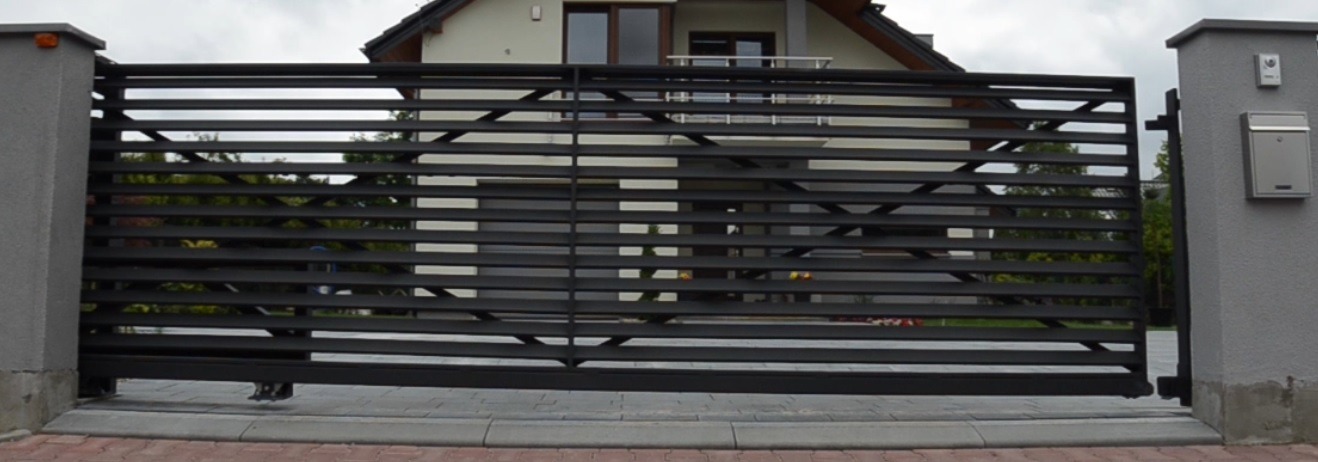 Nowoczesna, przesuwna brama wjazdowa z poziomych, czarnych profili, zamontowana na betonowych słupkach, z widocznym domem w tle.