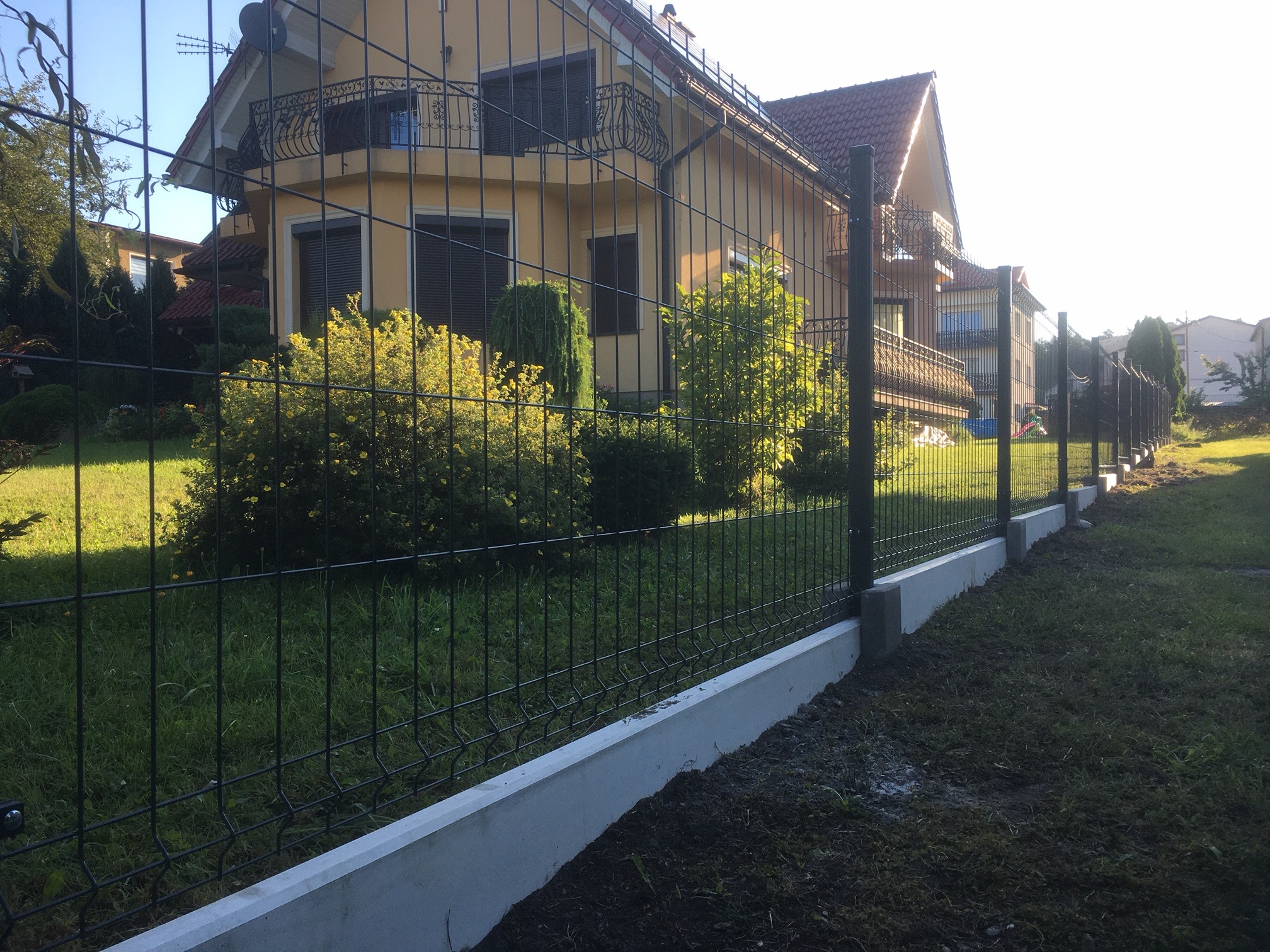 Czarny panelowy płot na betonowej podmurówce, widok z bliska na tle dwupiętrowego domu z balkonem, zieleń w tle.