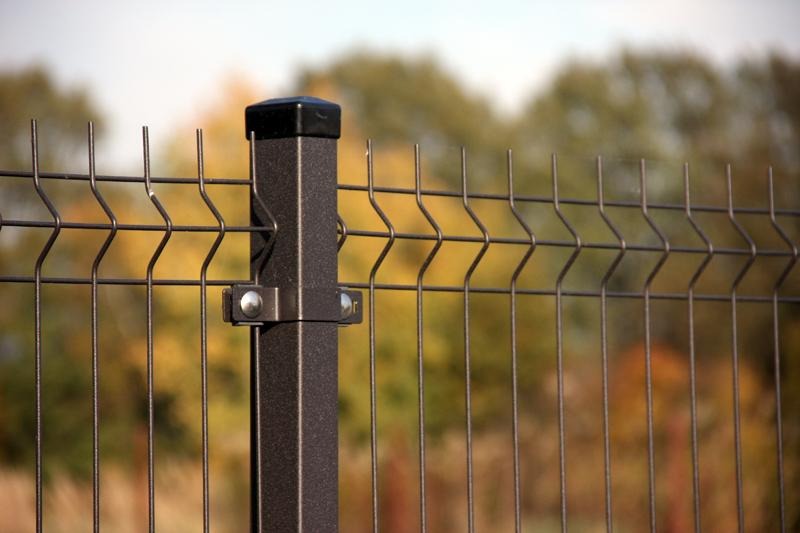 Fragment brązowego ogrodzenia panelowego z metalowym słupkiem i nasadką, widok z bliska na tle jesiennego krajobrazu.