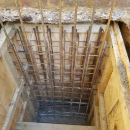 Zbrojenie pionowe i poziome w drewnianym szalunku przygotowane pod wylewkę betonową fundamentu. Widoczne wystające pręty zbrojeniowe oraz fragment starego muru ceglanego.