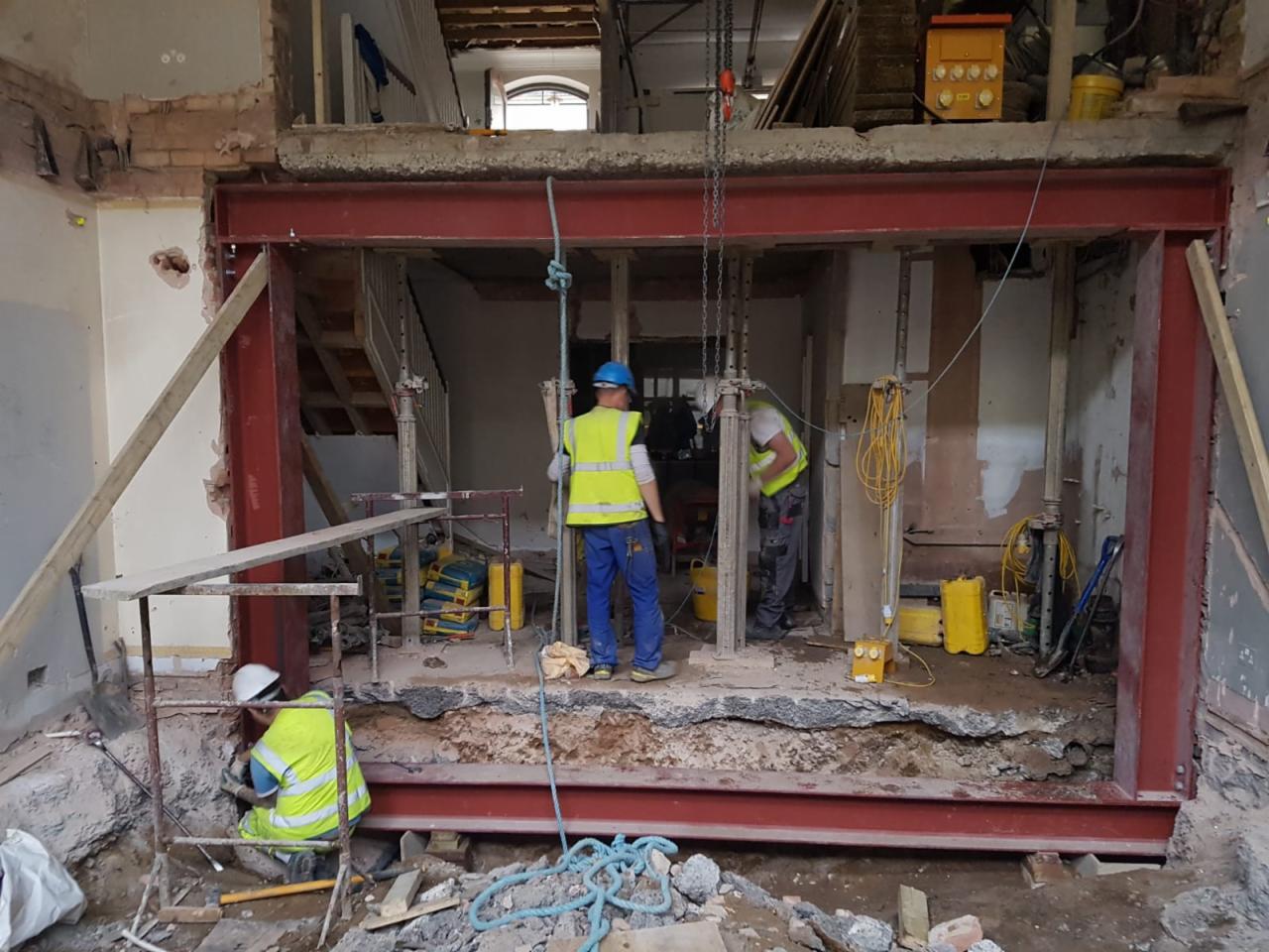 Pracownicy w kaskach i kamizelkach odblaskowych montują stalową konstrukcję nośną w trakcie remontu wnętrza, widoczne narzędzia i materiały budowlane.