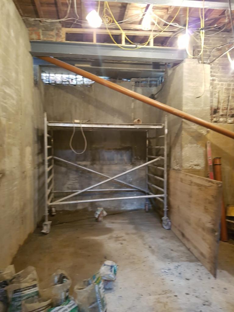 Wnętrze w trakcie remontu: surowe ściany, metalowa konstrukcja pod sufitem, rusztowanie na kółkach, worki z materiałami budowlanymi na podłodze, tymczasowe oświetlenie.