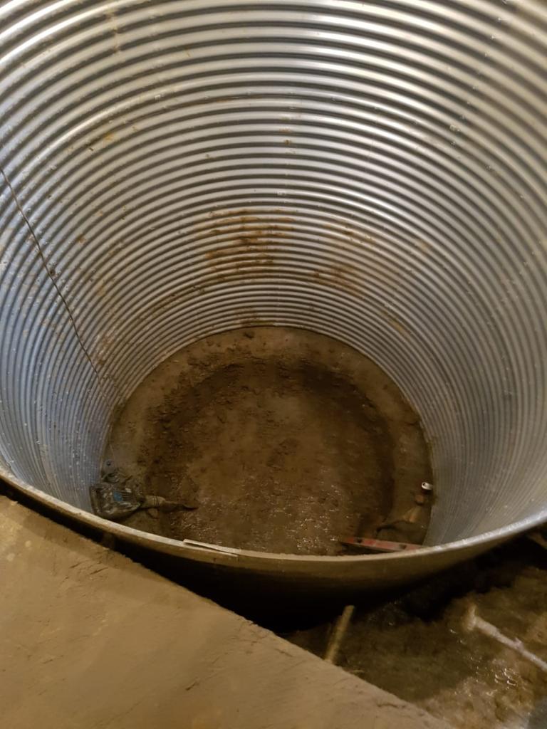 Wnętrze dużej, metalowej rury falistej z ziemią na dnie, widoczne narzędzia budowlane, potencjalnie element konstrukcyjny studni lub zbiornika podziemnego.