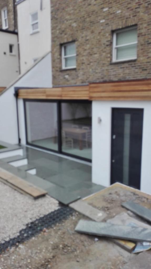 Nowoczesna dobudówka z dużym przesuwnym oknem i ciemnymi drzwiami wejściowymi, wykończona drewnem na zewnątrz, widoczne prace wykończeniowe wokół.