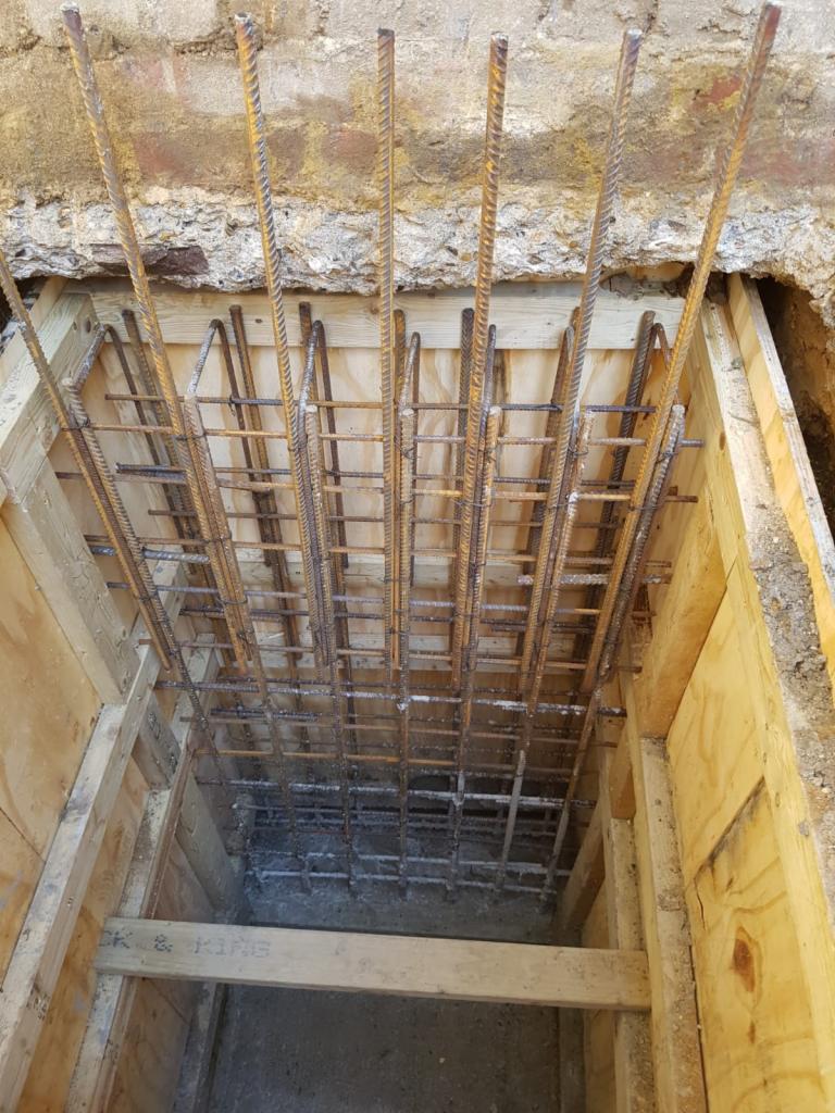 Zbrojenie pionowe i poziome w drewnianym szalunku przygotowane pod wylewkę betonową fundamentu. Widoczne wystające pręty zbrojeniowe oraz fragment starego muru ceglanego.