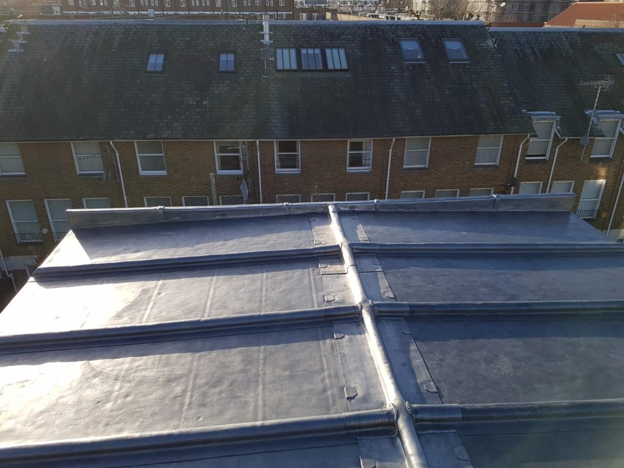 Szary dach pokryty blachą z widocznymi łączeniami na tle budynku mieszkalnego z cegły.