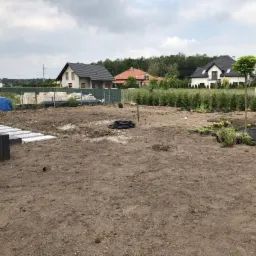 Przygotowanie terenu pod ogród: wyrównana ziemia, ułożone obrzeża z kostki, sadzenie roślin w czarnej agrowłókninie, widoczne drzewa i domy w tle.