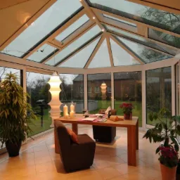 Szklana oranżeria z białymi ramami, widok na ogród, stół z nakryciem i świecami, rośliny doniczkowe wewnątrz.