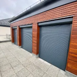 Trzy szare, metalowe osłony okienne w budynku obłożonym drewnem, widok z zewnątrz na szarym brukowanym tarasie.