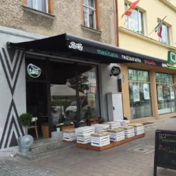 Zewnętrzny widok restauracji z czarną markizą z napisem 'Pepsi-Cola' i 'mexicano restaurante pequeño', neonowym logo 'tako', wejściem ozdobionym geometrycznym wzorem i skrzynkami służącymi jako...