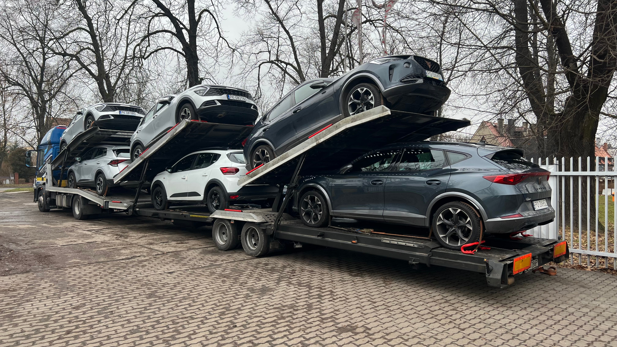 Wielopoziomowa laweta załadowana różnymi modelami samochodów osobowych, zabezpieczonych pasami transportowymi, na tle szarej kostki brukowej i drzew bez liści.