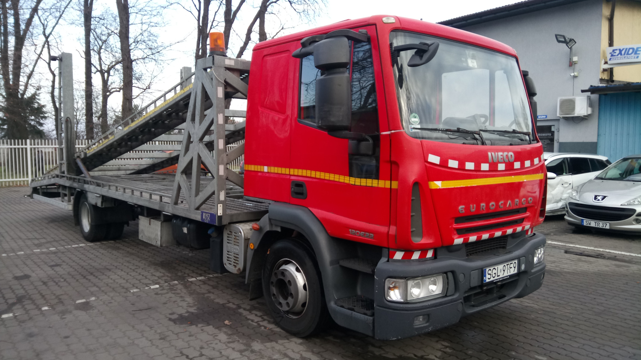 Czerwona laweta marki Iveco Eurocargo z podniesioną platformą, zaparkowana na brukowanym placu, z uszkodzonym białym samochodem i srebrnym Peugeotem w tle.