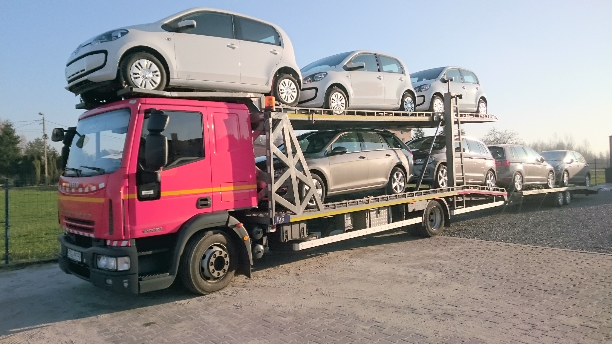 Różowy samochód ciężarowy z lawetą przewożący sześć szarych samochodów osobowych na dwupoziomowej platformie transportowej.
