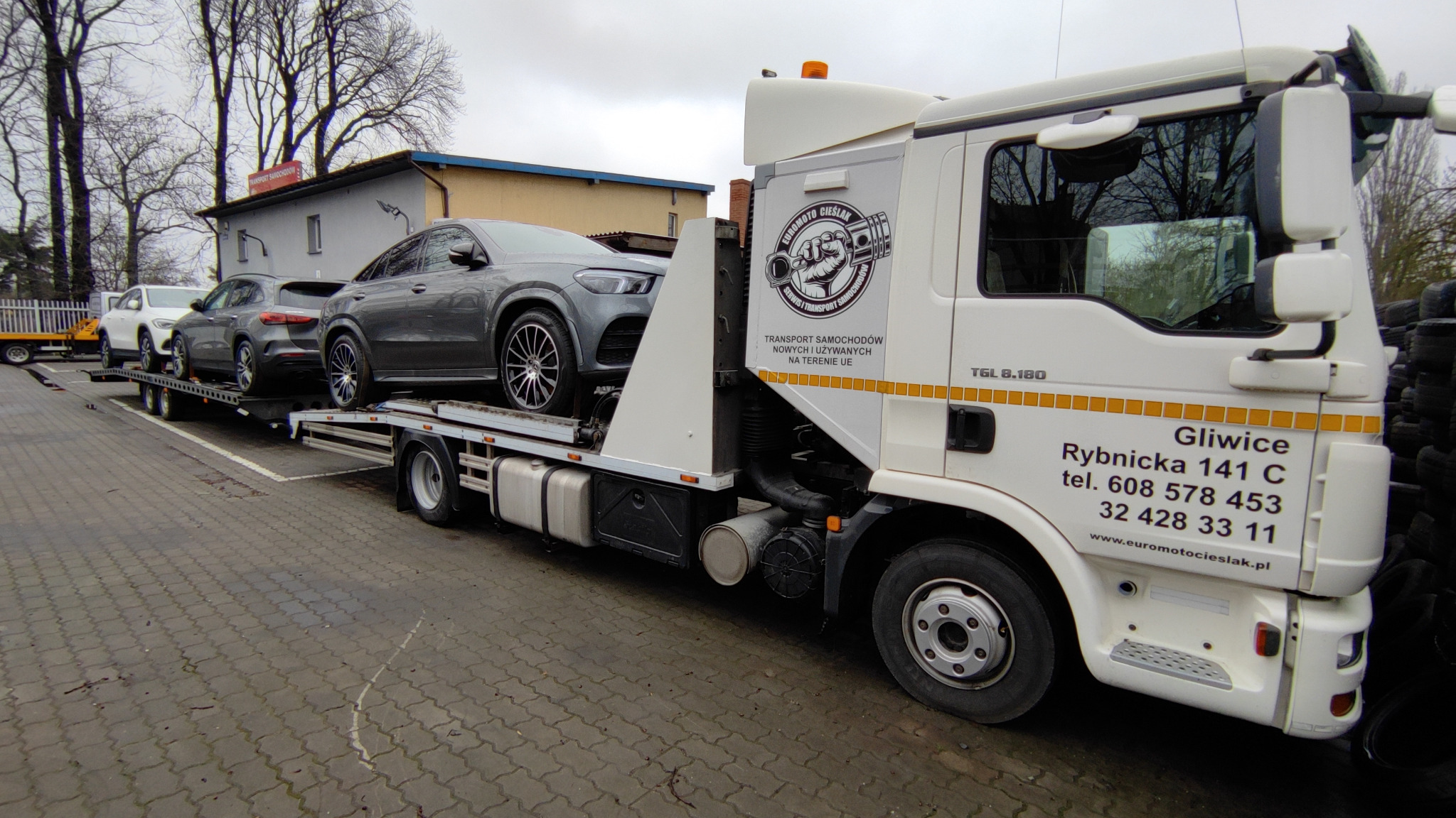 Biała laweta MAN TGL 8.180 z trzema szarymi samochodami osobowymi marki Mercedes na platformie, zaparkowana na kostce brukowej, widoczny budynek i drzewa w tle, logo firmy transportowej na kabinie