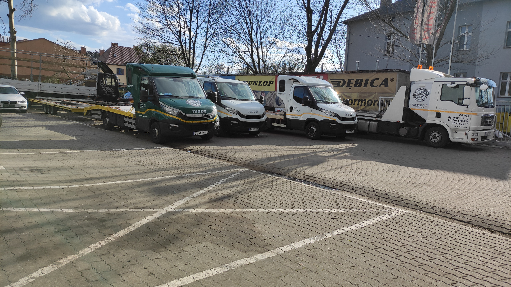 Flota pojazdów lawet i ciężarówek Iveco oraz MAN zaparkowana na placu z kostki brukowej w słoneczny dzień, widoczne reklamy opon Dunlop i Dębica na naczepach, budynki w tle.