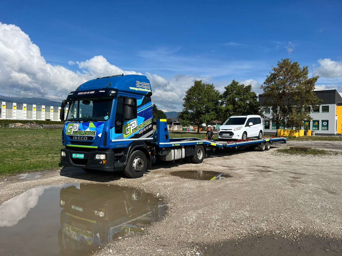 Niebieska laweta Iveco z logo GP Automotive przewozi białego Forda Transita na tle błękitnego nieba i zieleni, odbicie pojazdu w kałuży na żwirowym podłożu.