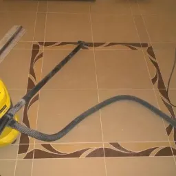 Beżowe płytki podłogowe z dekoracyjnym brązowym obramowaniem w trakcie czyszczenia odkurzaczem przemysłowym Karcher, obok listwa przypodłogowa.