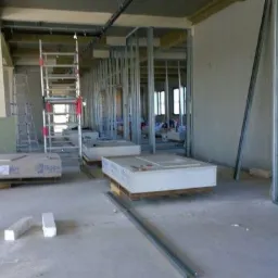 Wnętrze w trakcie szpachlowania: metalowe profile ścian działowych, płyty gipsowo-kartonowe na paletach, poziomica oparta o ścianę, drabina aluminiowa.