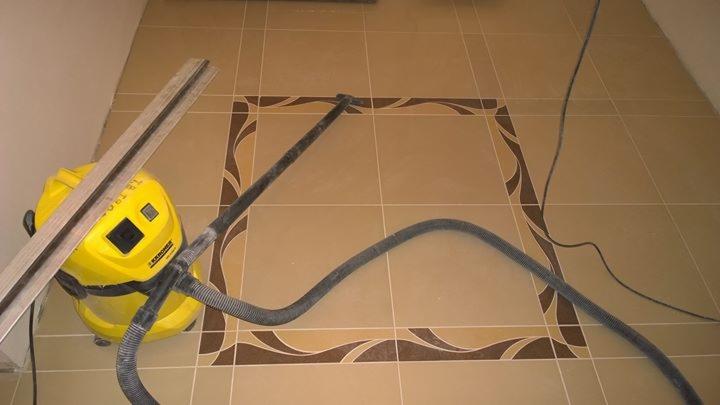 Beżowe płytki podłogowe z dekoracyjnym brązowym obramowaniem w trakcie czyszczenia odkurzaczem przemysłowym Karcher, obok listwa przypodłogowa.