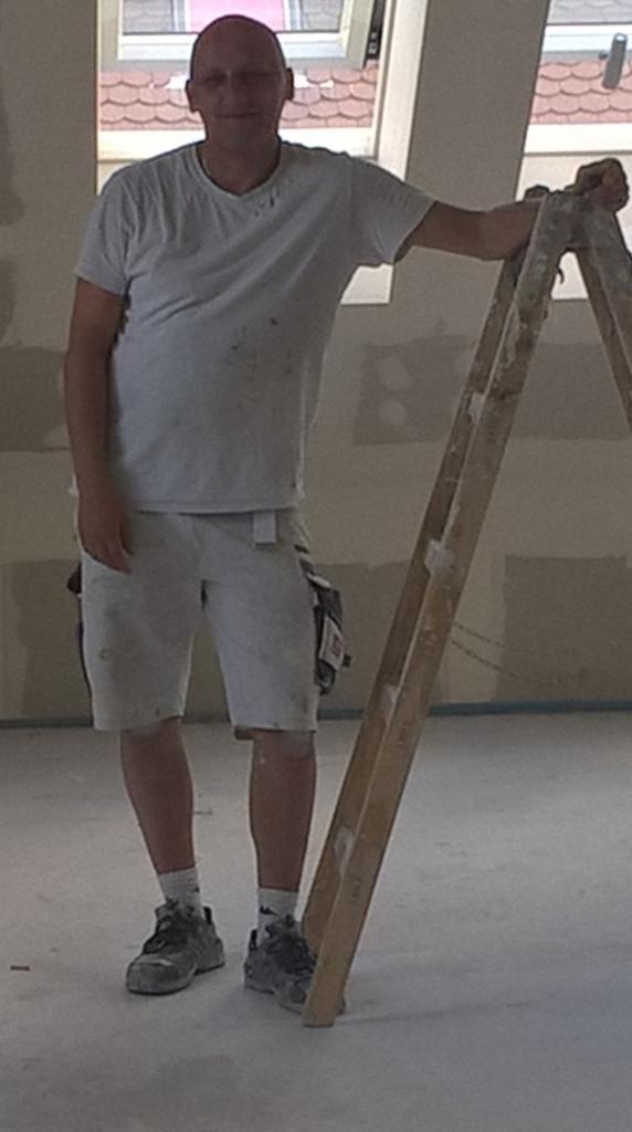 Mężczyzna w ubraniu roboczym opiera się o drewnianą drabinę w pomieszczeniu ze ścianami przygotowanymi do malowania, widoczne okna dachowe.