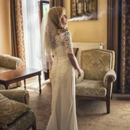 Panna młoda w białej sukni z koronkowymi rękawami i welonem, stojąca tyłem, z tatuażem na ramieniu, paląca papierosa w pokoju z klasycznymi meblami i obrazem na ścianie.