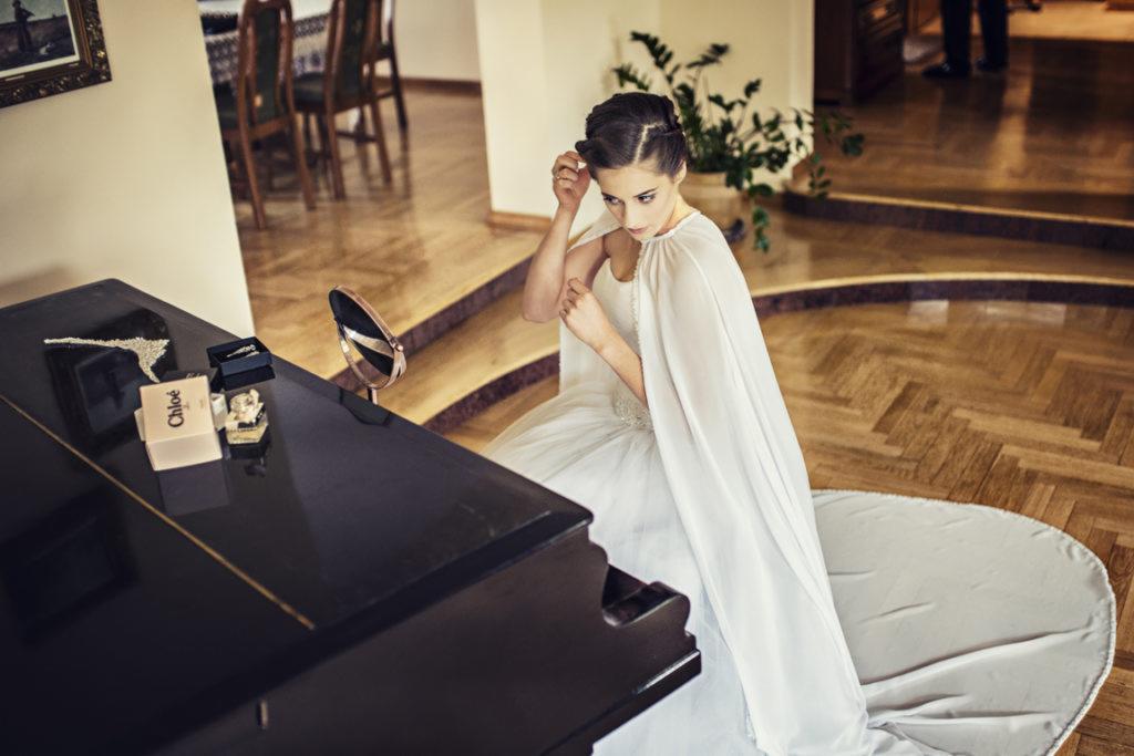 Panna młoda w białej sukni z peleryną, poprawia włosy przed lustrem stojącym na fortepianie, obok leżą pudełka z biżuterią, drewniana podłoga w tle.