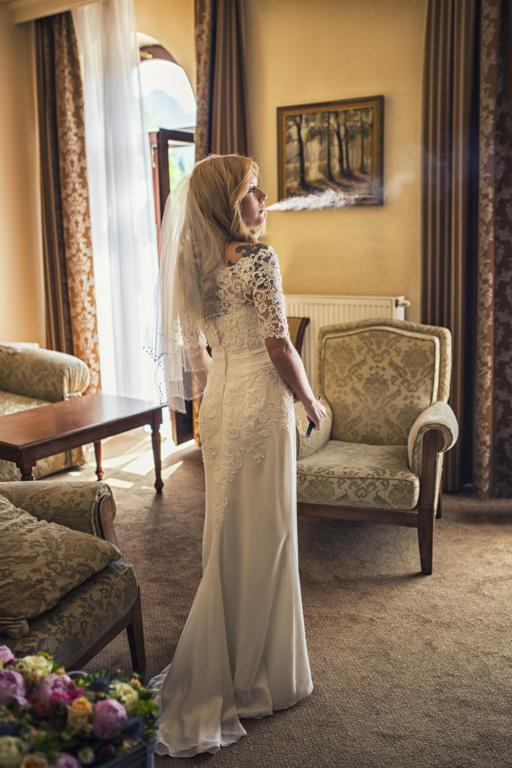 Panna młoda w białej sukni z koronkowymi rękawami i welonem, stojąca tyłem, z tatuażem na ramieniu, paląca papierosa w pokoju z klasycznymi meblami i obrazem na ścianie.