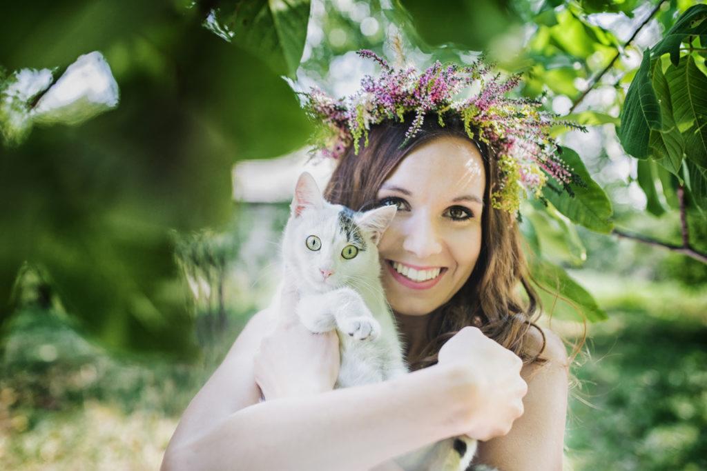 Uśmiechnięta kobieta z wiankiem z polnych kwiatów trzymająca białego kota z szarymi łatami na tle zieleni.