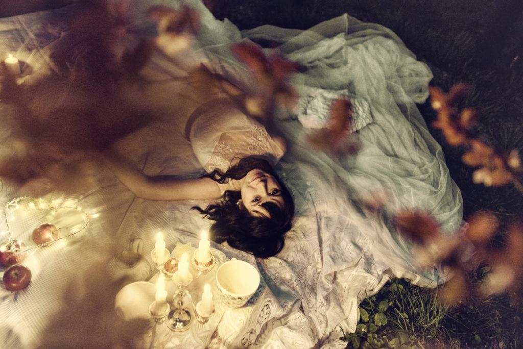 Młoda kobieta w koronkowej sukience leży na kocu w plenerze, otoczona świecami, jabłkami i drobnymi lampkami, ujęcie z góry.