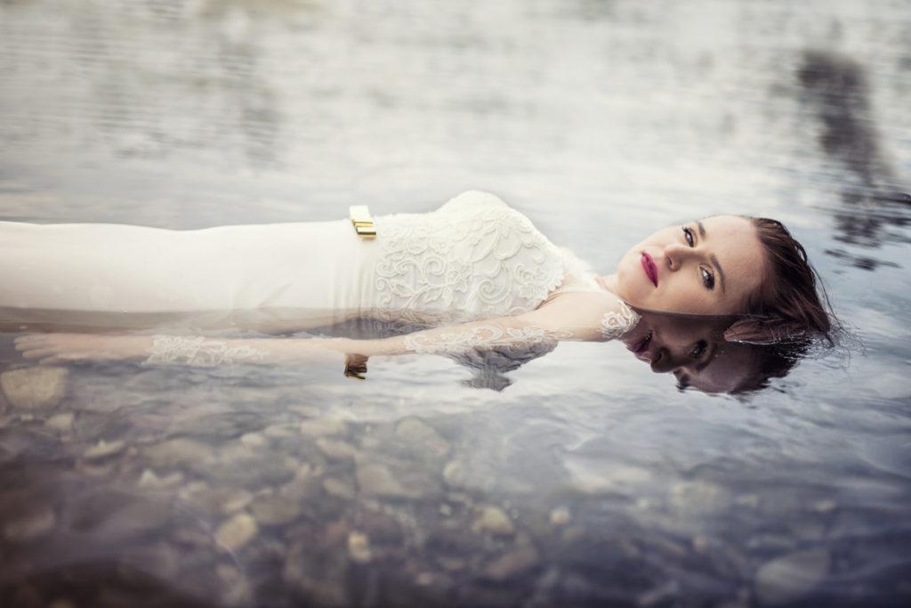 Panna młoda w białej sukni z koronkowymi rękawami i złotym paskiem, zanurzona w wodzie jeziora, odbicie twarzy na powierzchni wody.