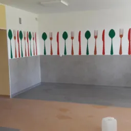 Wnętrze sali z nową, dwukolorową podłogą (szary i beżowy) oraz dekoracją ścienną przedstawiającą zielone łyżki i czerwone sztućce, widoczne drzwi ewakuacyjne.