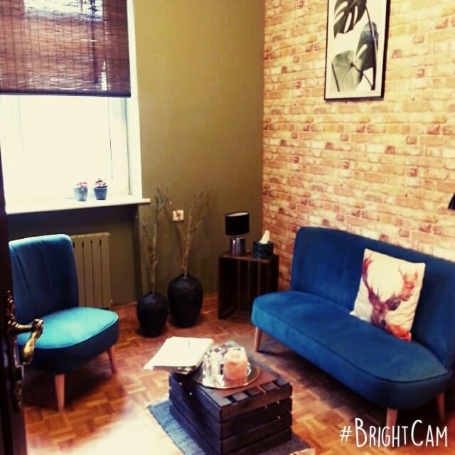 Przytulny gabinet z ceglaną ścianą, turkusowym fotelem i kanapą, drewnianą podłogą w jodełkę, stolikiem z książkami i dekoracjami, oraz roślinnym obrazem na ścianie.